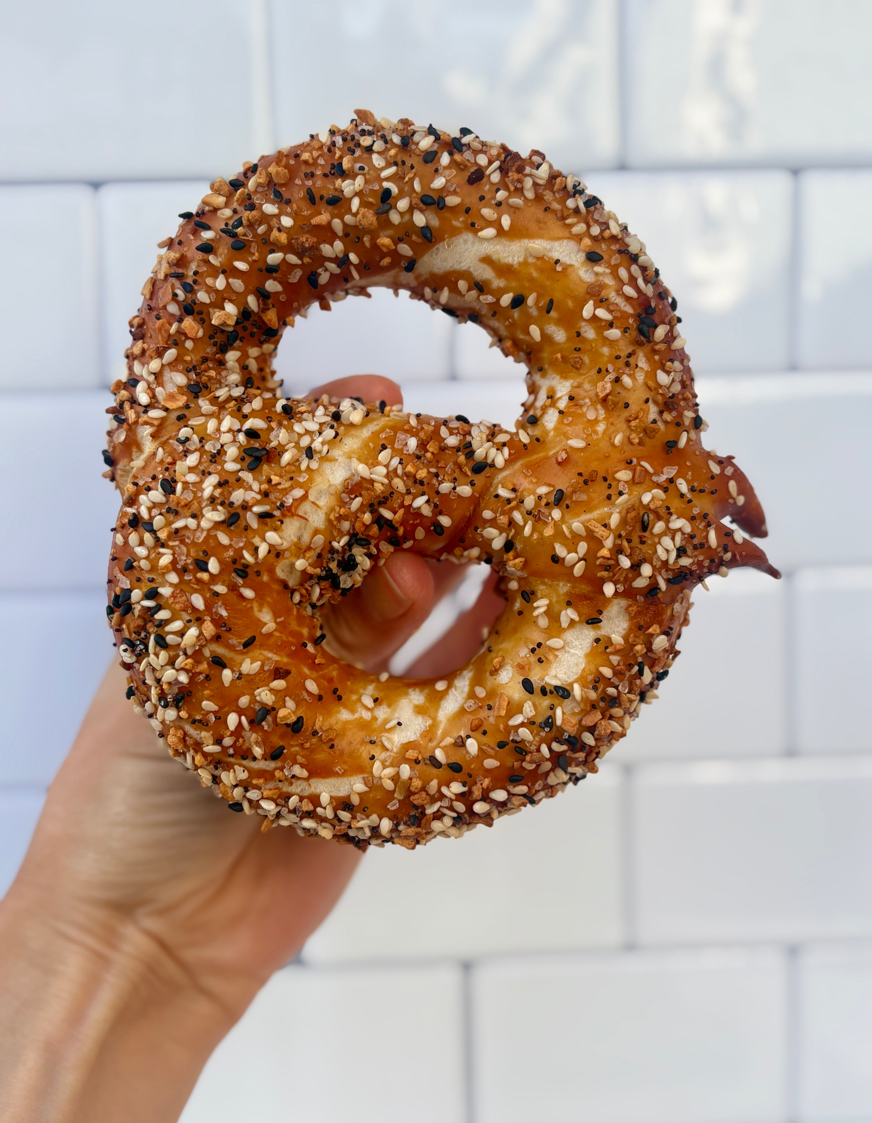 Hand holding a bagel with sesame seeds and poppy seeds against a white tiled wall.