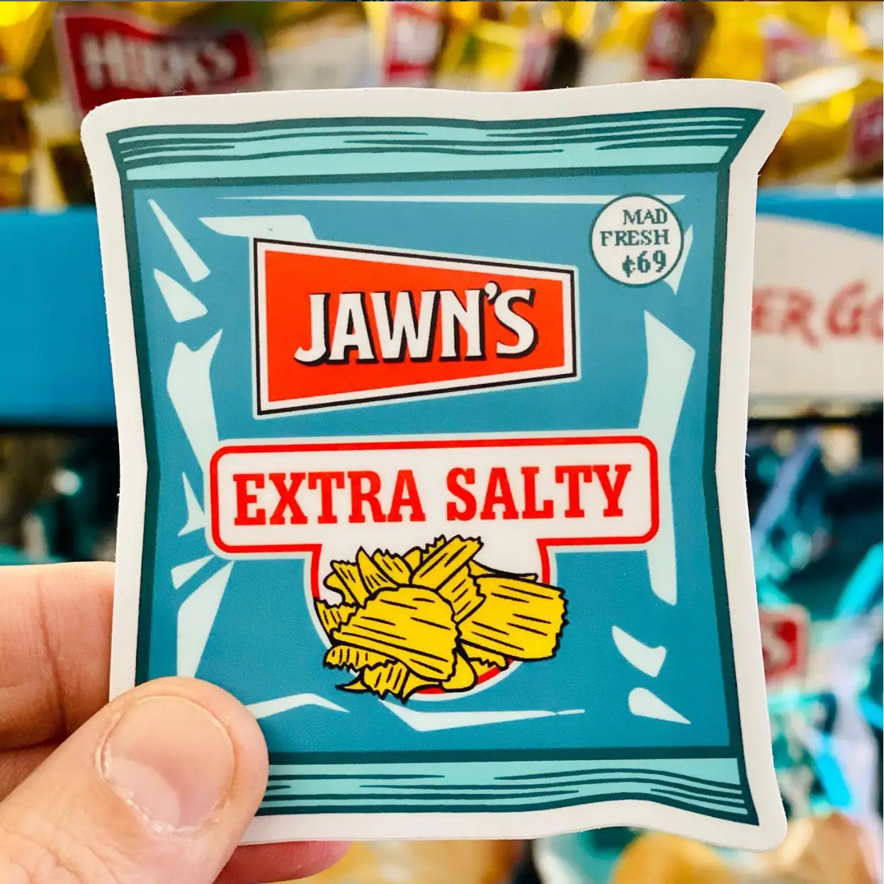Sticker of Jawn's Extra Salty chips packaging held in front of a store shelf.