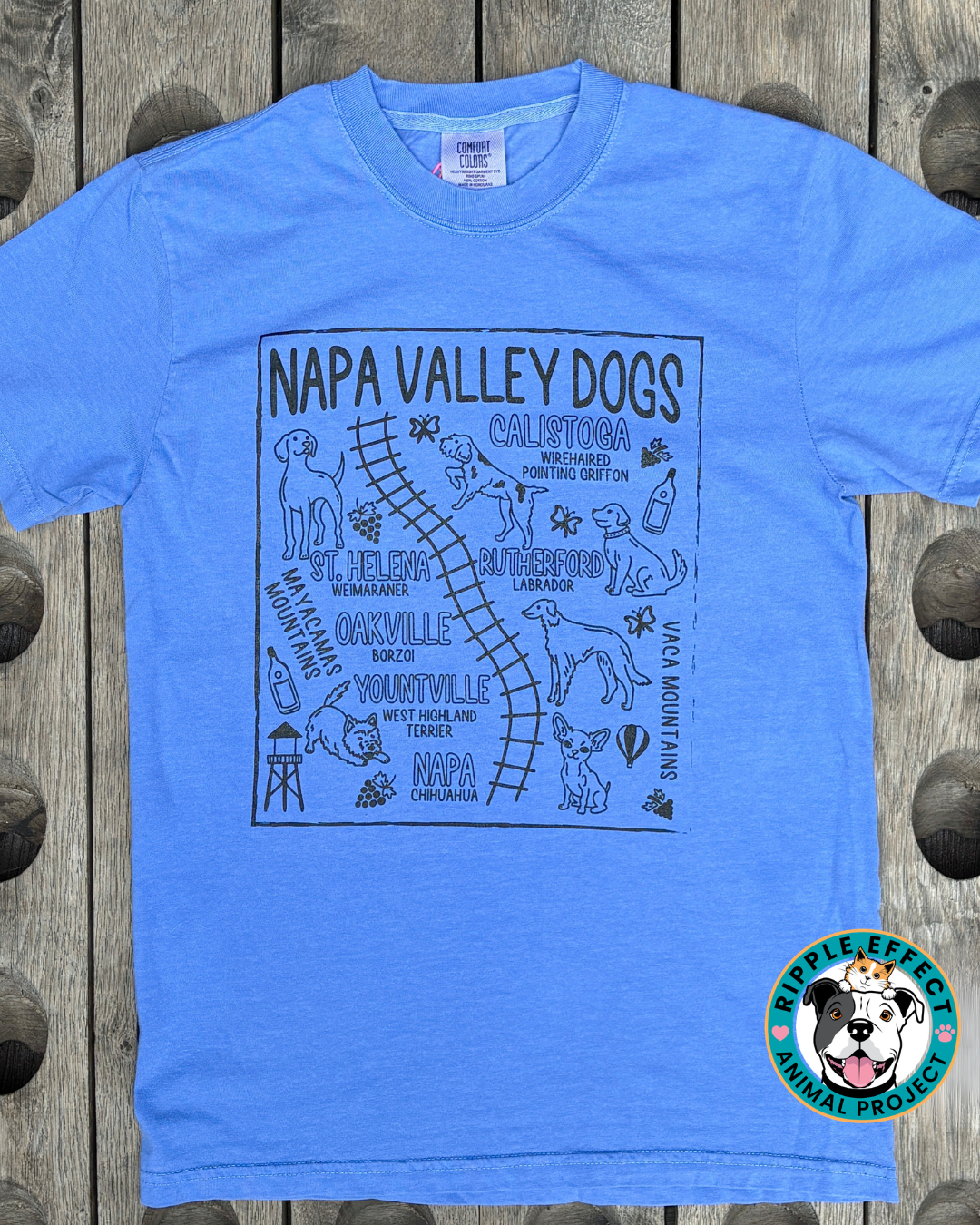 A light blue short-sleeve t-shirt with a black graphic print of a map labeled 'NAPA VALLEY DOGS' featuring dog illustrations and local landmarks. The shirt is laid flat on a rustic wooden background, with a 'Ripple Effects Animal Project' logo in the bottom right corner.
