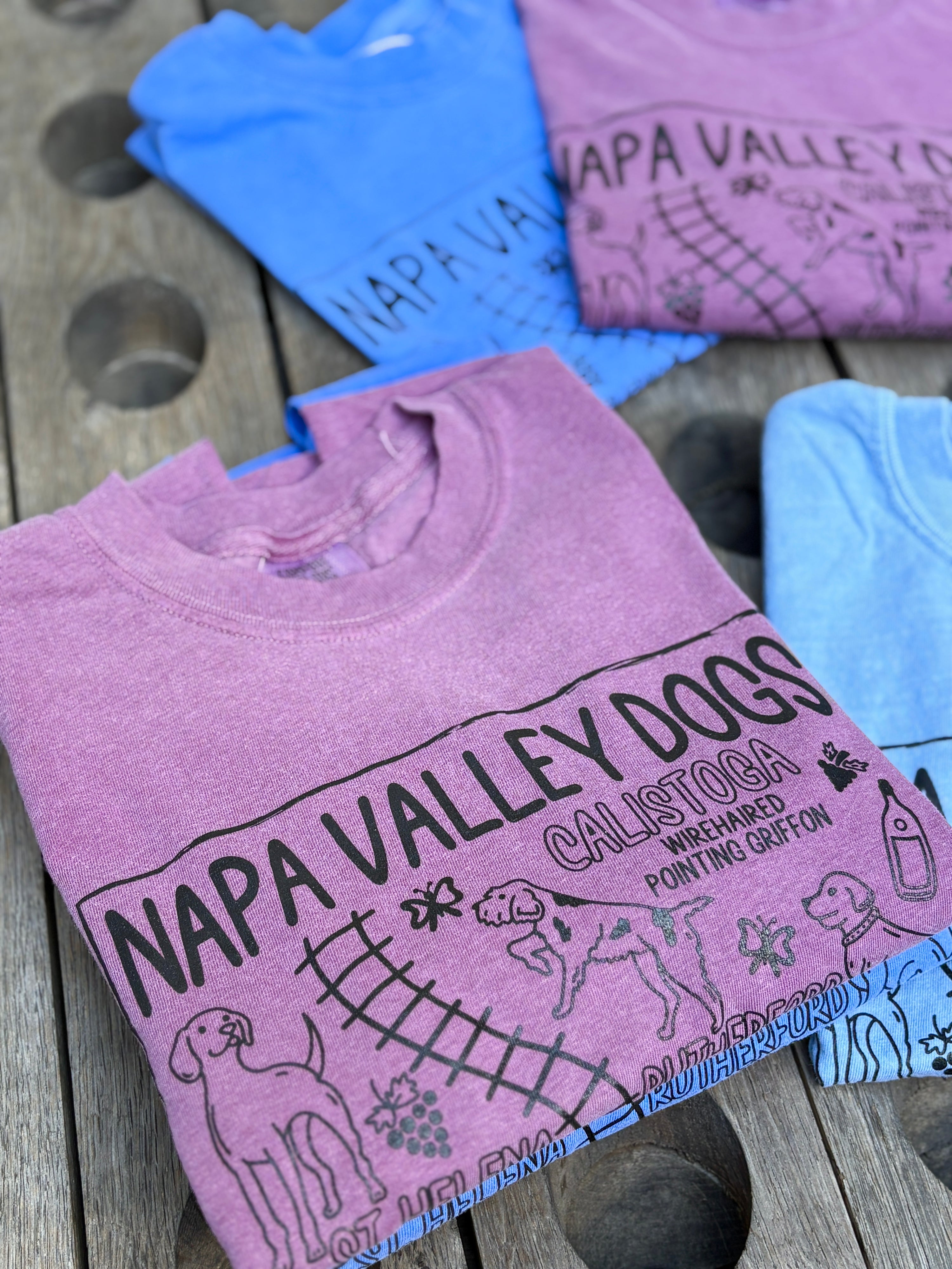 A folded heather pink graphic T-shirt on a rustic wooden surface, with other folded T-shirts partially visible behind it in blue and pink. The front shirt has black printed text reading 'NAPA VALLEY DOGS' and 'CALISTOGA WHEATHEADS POINTING GRIFTON' with dog and basket illustrations.