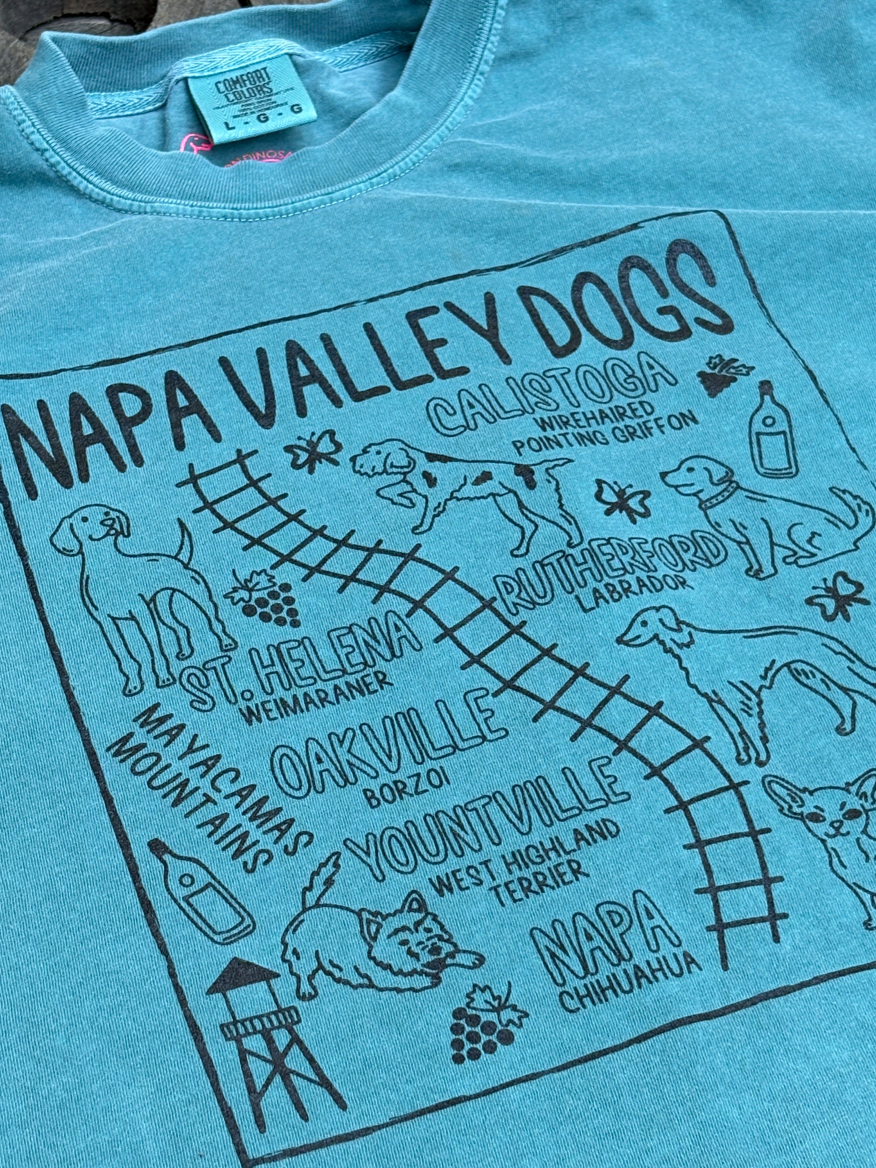 A close-up of a light blue graphic t-shirt featuring black printed text and illustrations for 'Napa Valley Dogs' with dog and chihuahua drawings and place names.