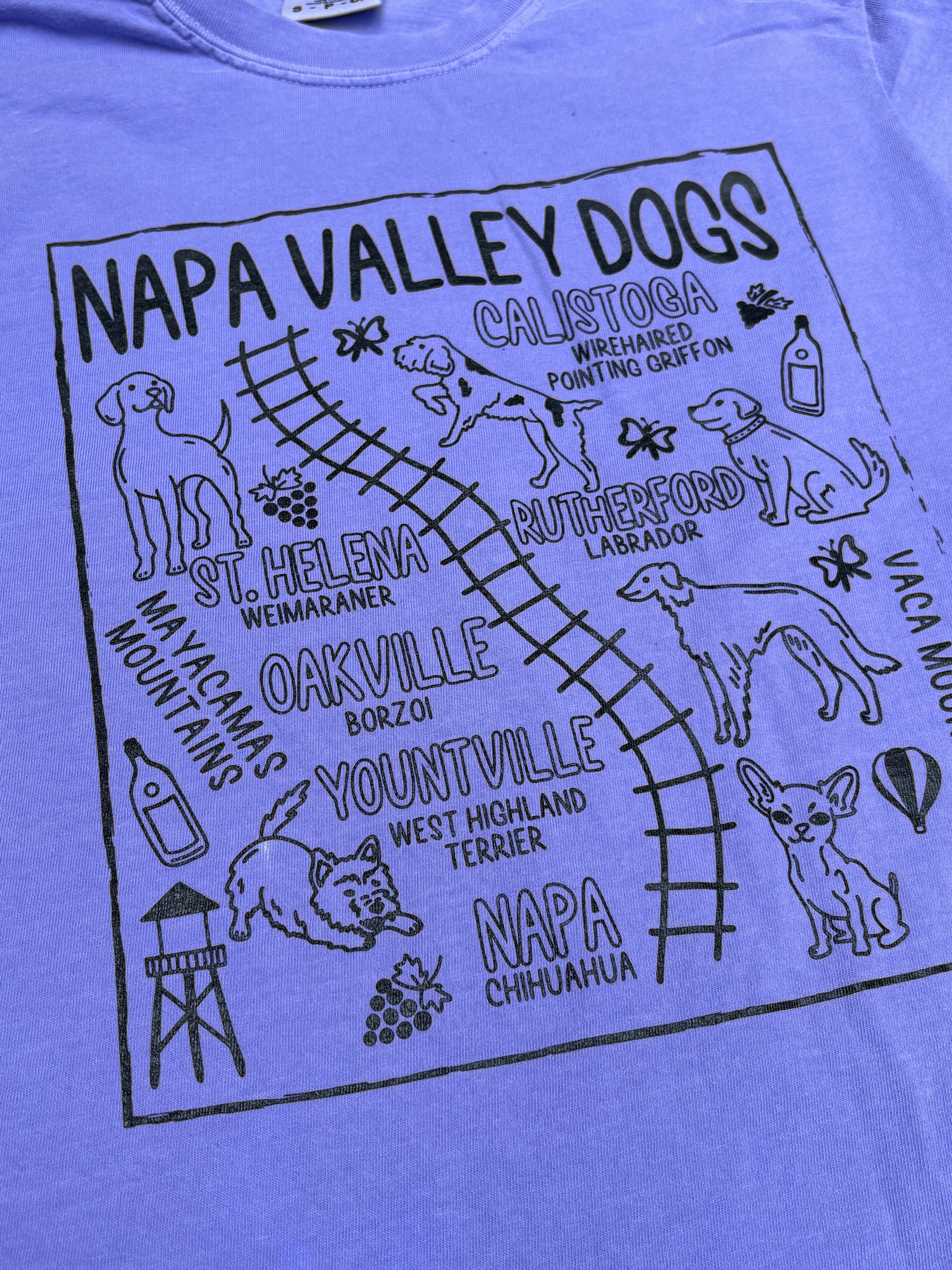 A close-up of a lavender purple t-shirt featuring a black graphic print titled 'NAPA VALLEY DOGS' with various dog illustrations and local place names.