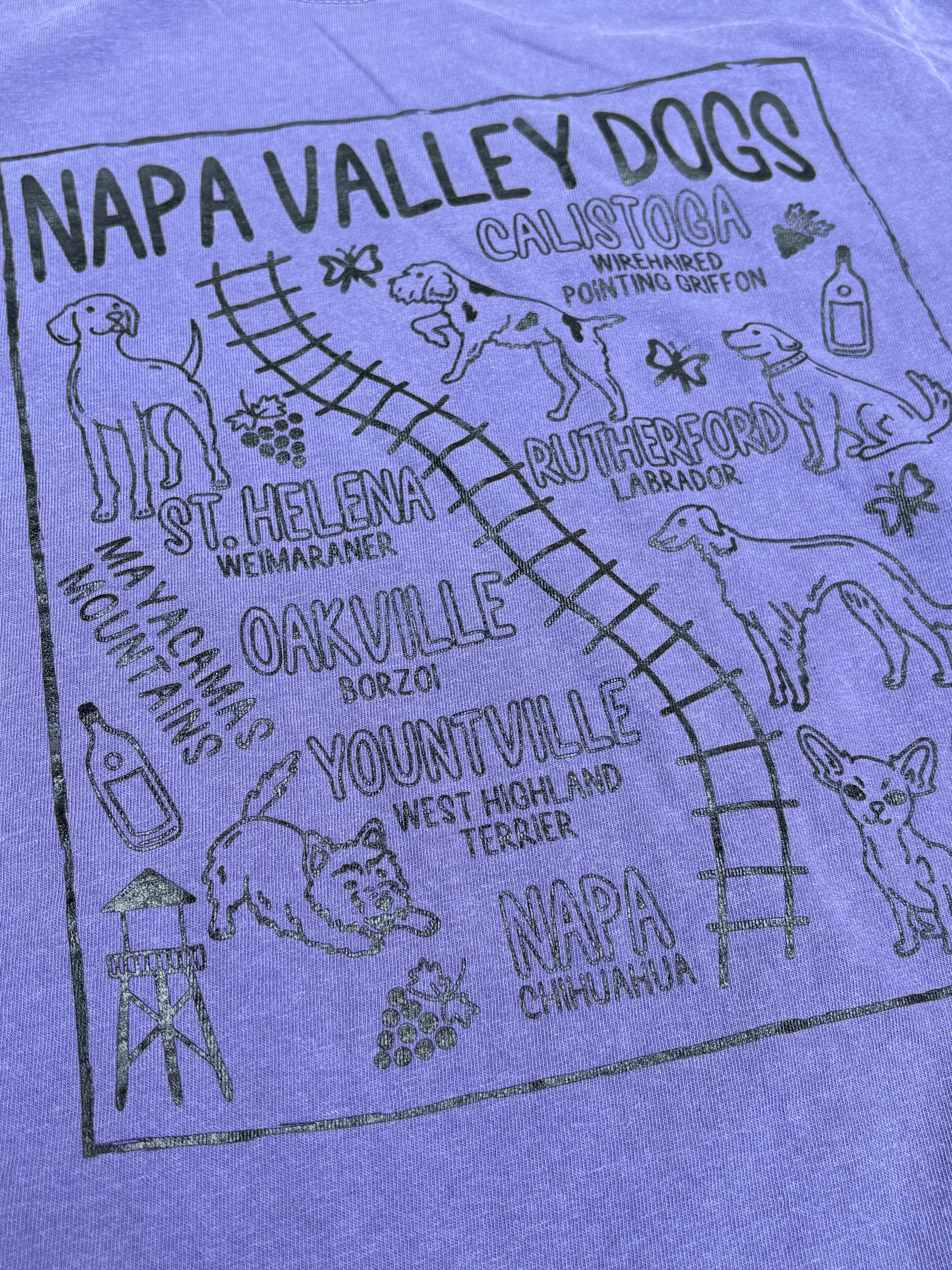 A close-up of a heather purple graphic t-shirt featuring a black printed design for 'Napa Valley Dogs' with illustrations of various dog breeds and local place names.