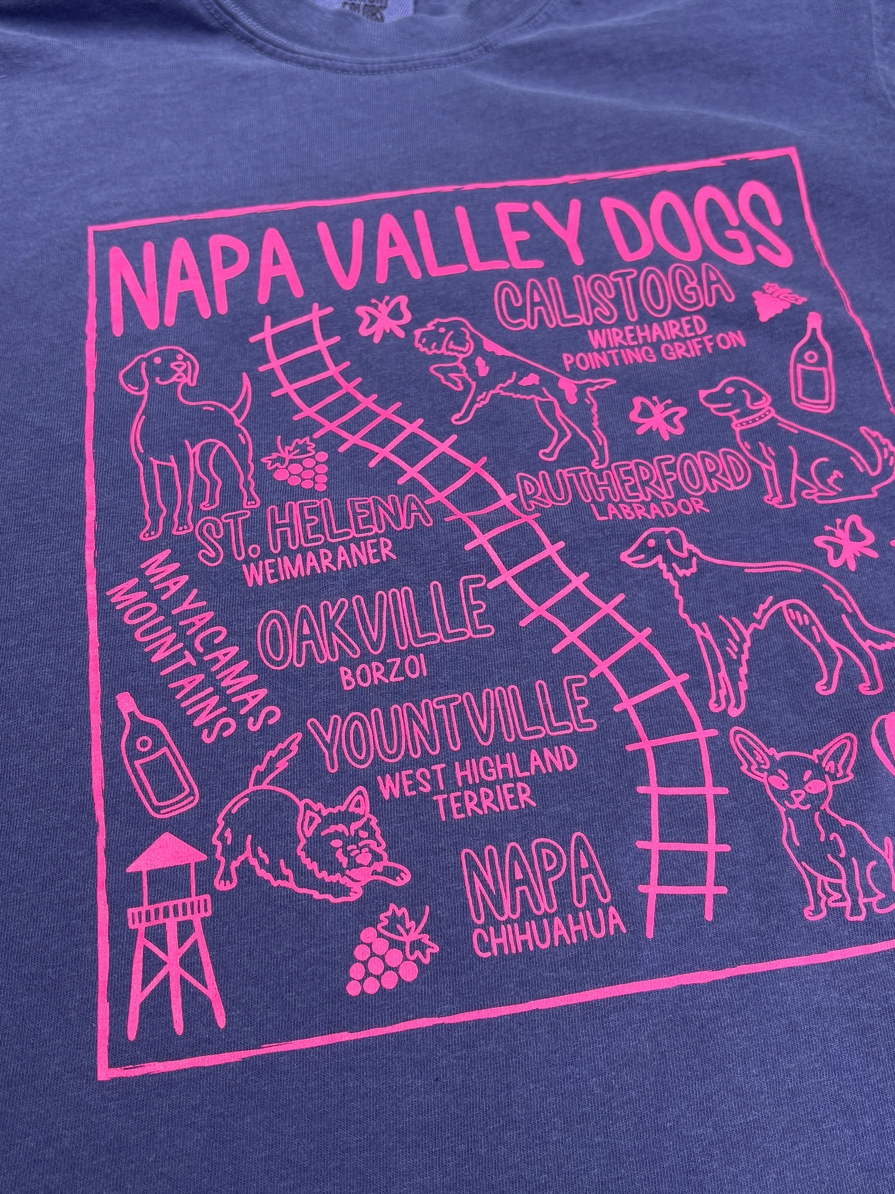 A close-up of a navy blue t-shirt featuring a bright pink graphic print with the text 'NAPA VALLEY DOGS' and various dog breeds, places, and illustrations.