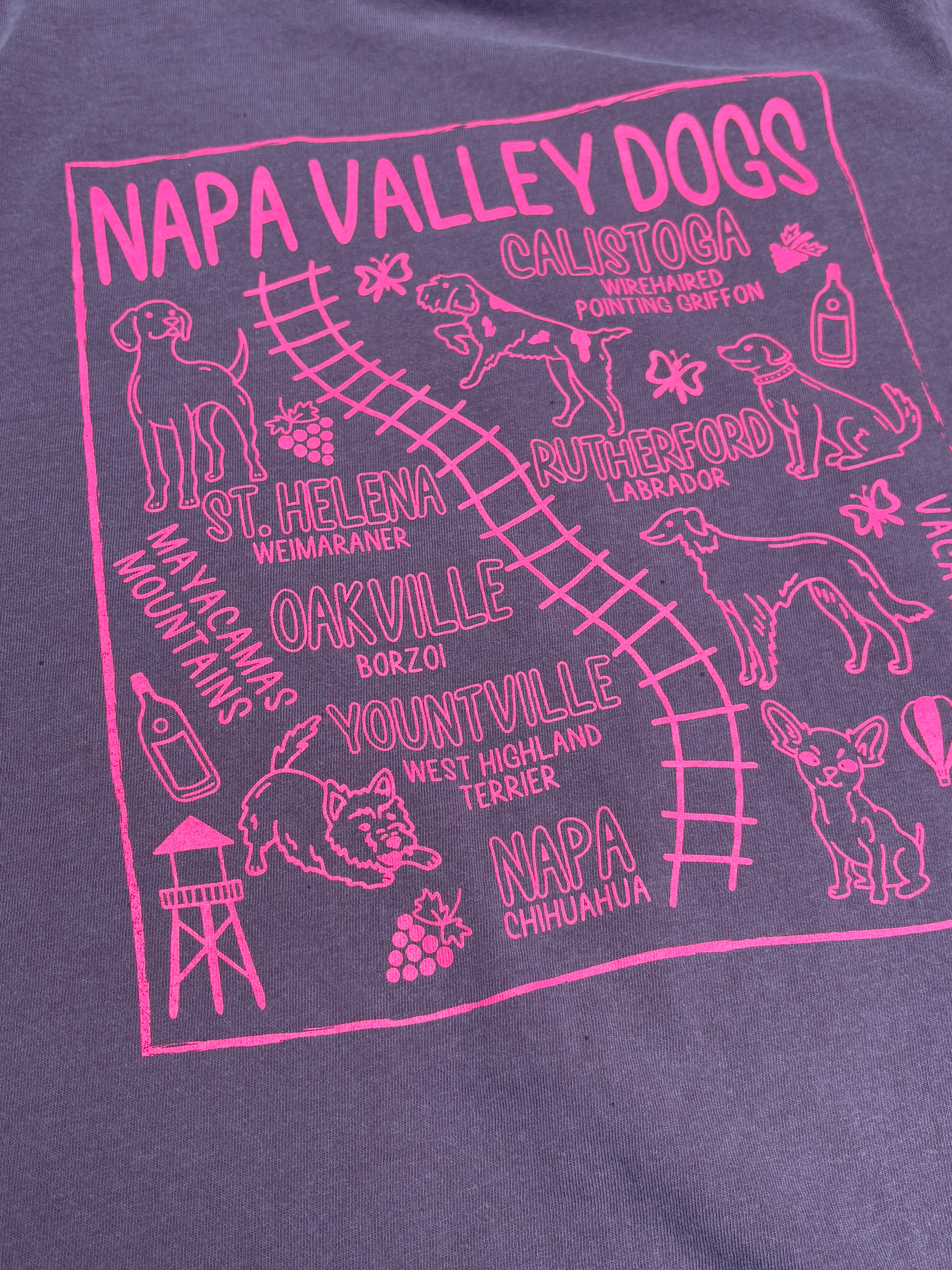 A close-up of a heathered purple t-shirt featuring a vibrant pink graphic print. The graphic is titled 'NAPA VALLEY DOGS' and displays various dog breeds, wine bottles, grapes, and local places like St.