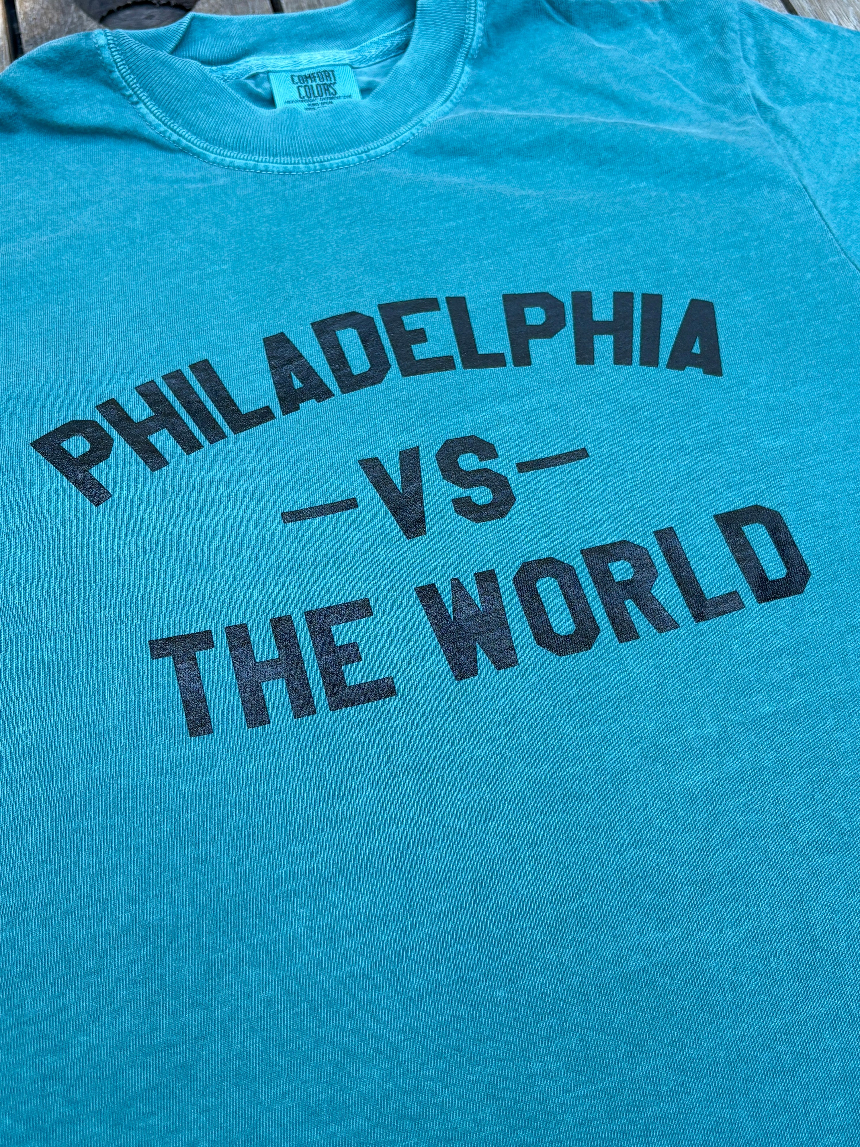 A close-up of a heathered turquoise blue t-shirt featuring the text 'PHILADELPHIA -VS- THE WORLD' printed in distressed black block letters across the chest.