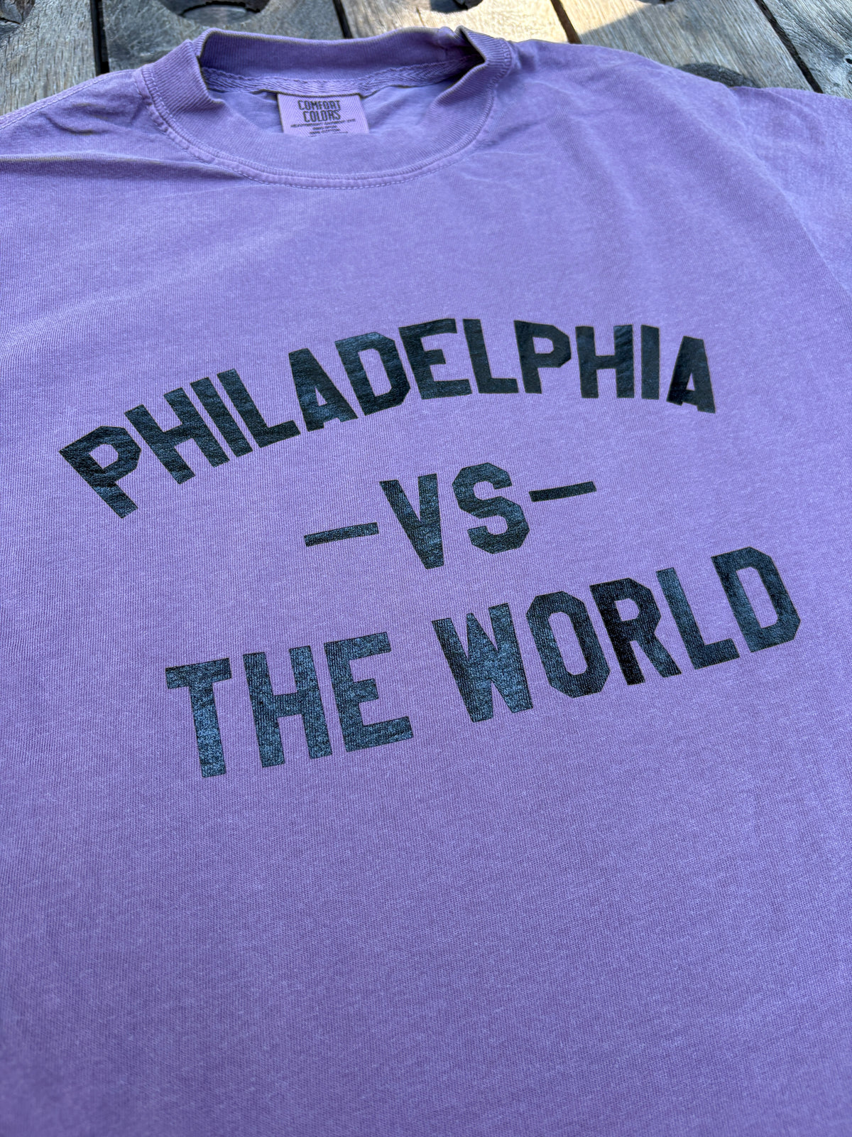A close-up of a lavender-purple t-shirt laid flat on a wooden surface, featuring distressed black text that reads 'PHILADELPHIA -VS- THE WORLD'.