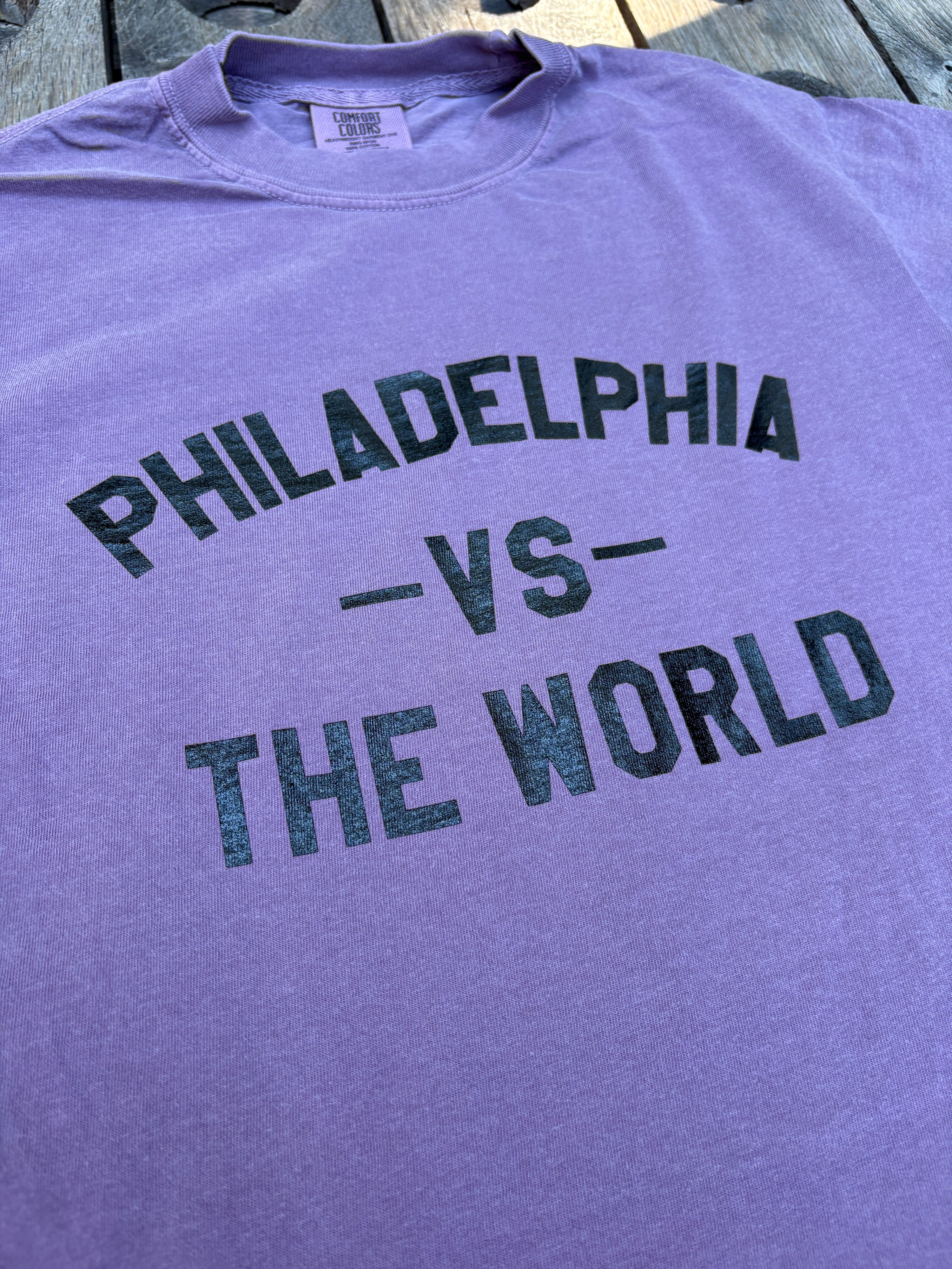A close-up of a lavender-purple t-shirt laid flat on a wooden surface, featuring distressed black text that reads 'PHILADELPHIA -VS- THE WORLD'.