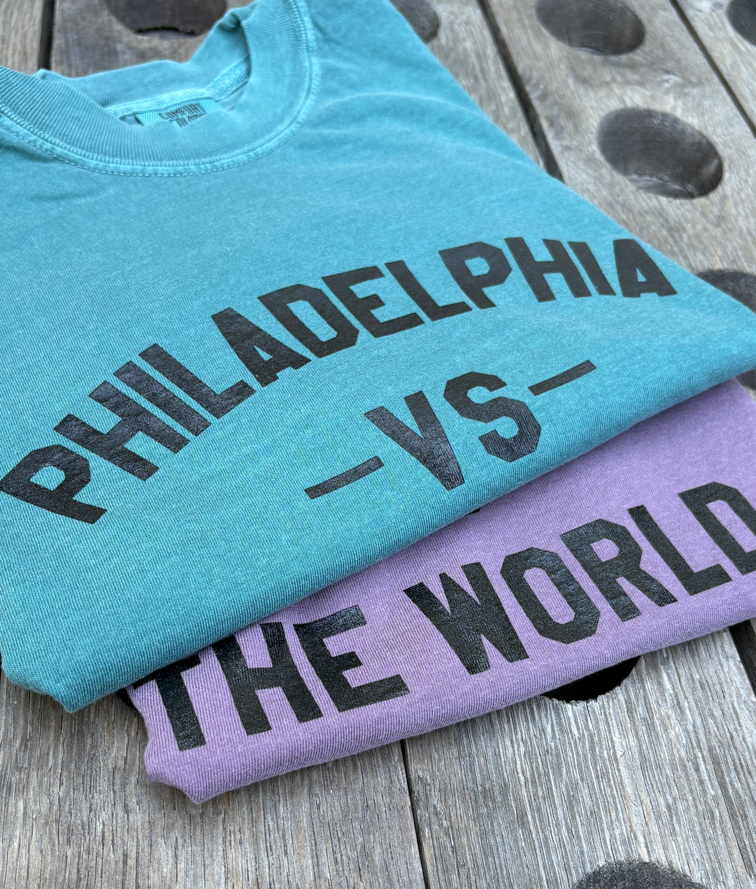 Two folded graphic T-shirts stacked on a weathered wooden surface. The top shirt is heather aqua with black text reading 'PHILADELPHIA -VS- THE WORLD', and the bottom shirt is heather lavender with black text reading 'THE WORLD'.