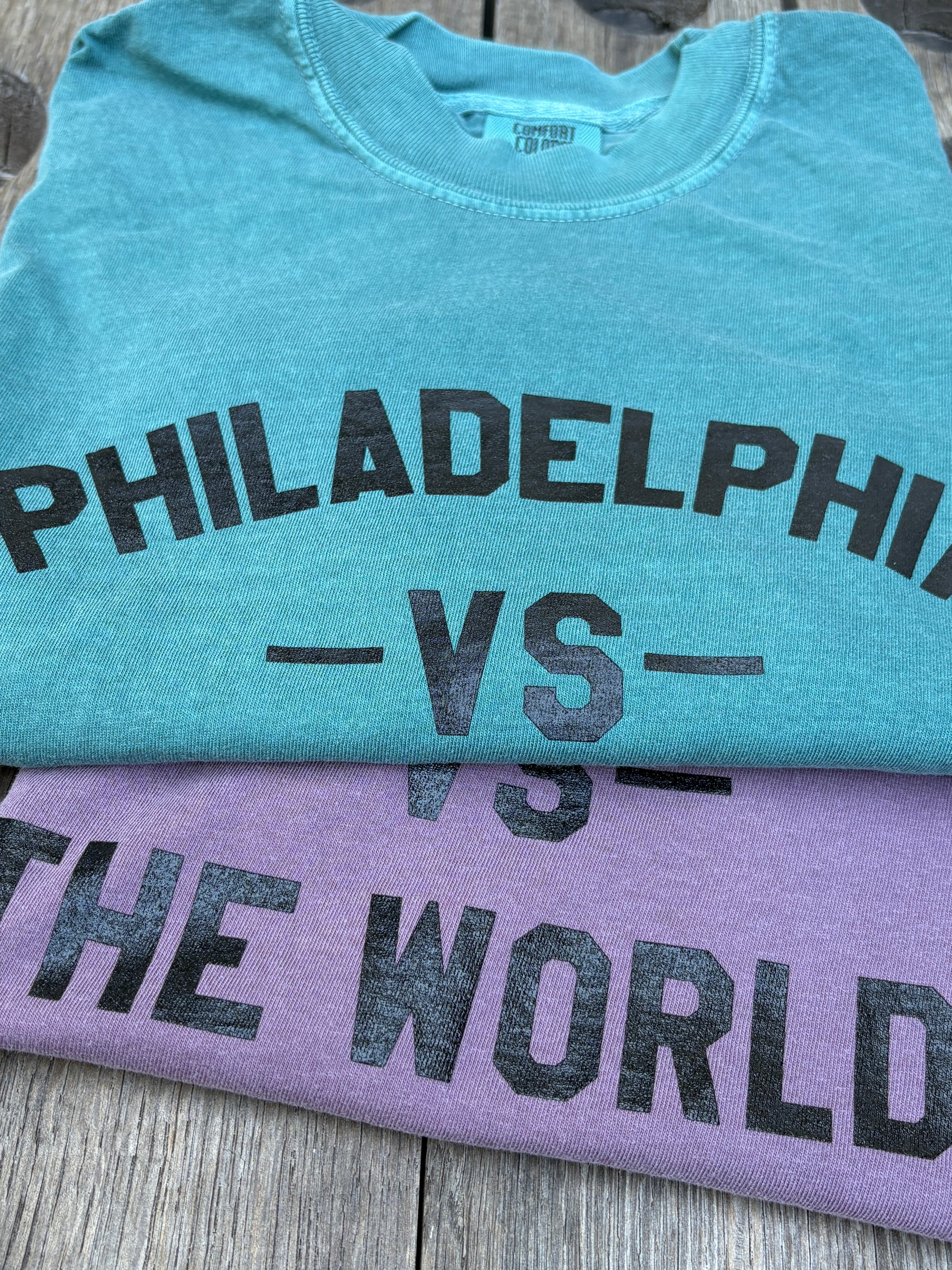 Two folded graphic T-shirts stacked on a weathered wood surface. The top shirt is teal with black text reading 'PHILADELPHIA -VS-', and the bottom shirt is lavender with black text reading 'THE WORLD'.