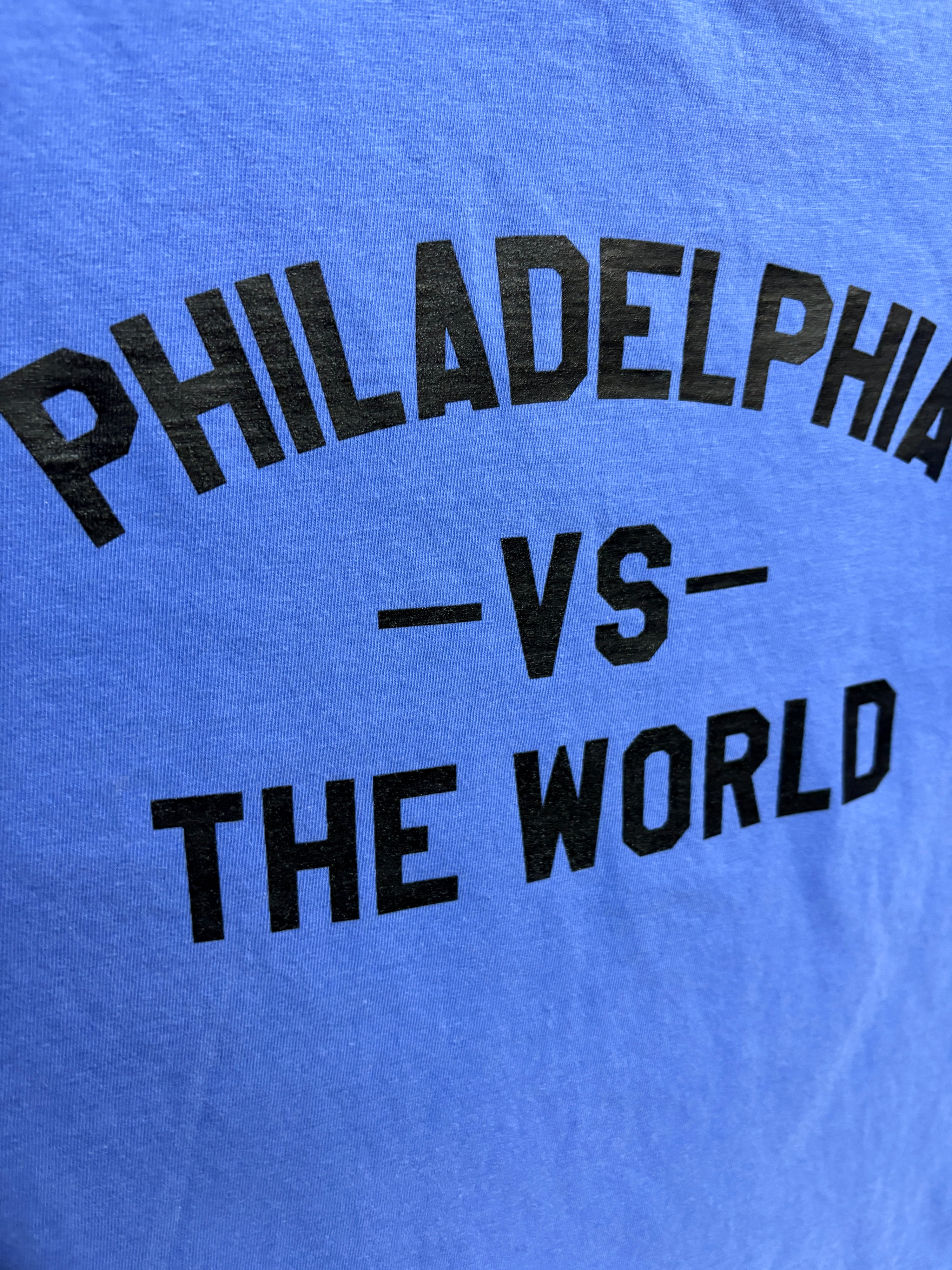 Close-up of a blue T-shirt with bold black text reading 'PHILADELPHIA -VS- THE WORLD' printed across the chest.