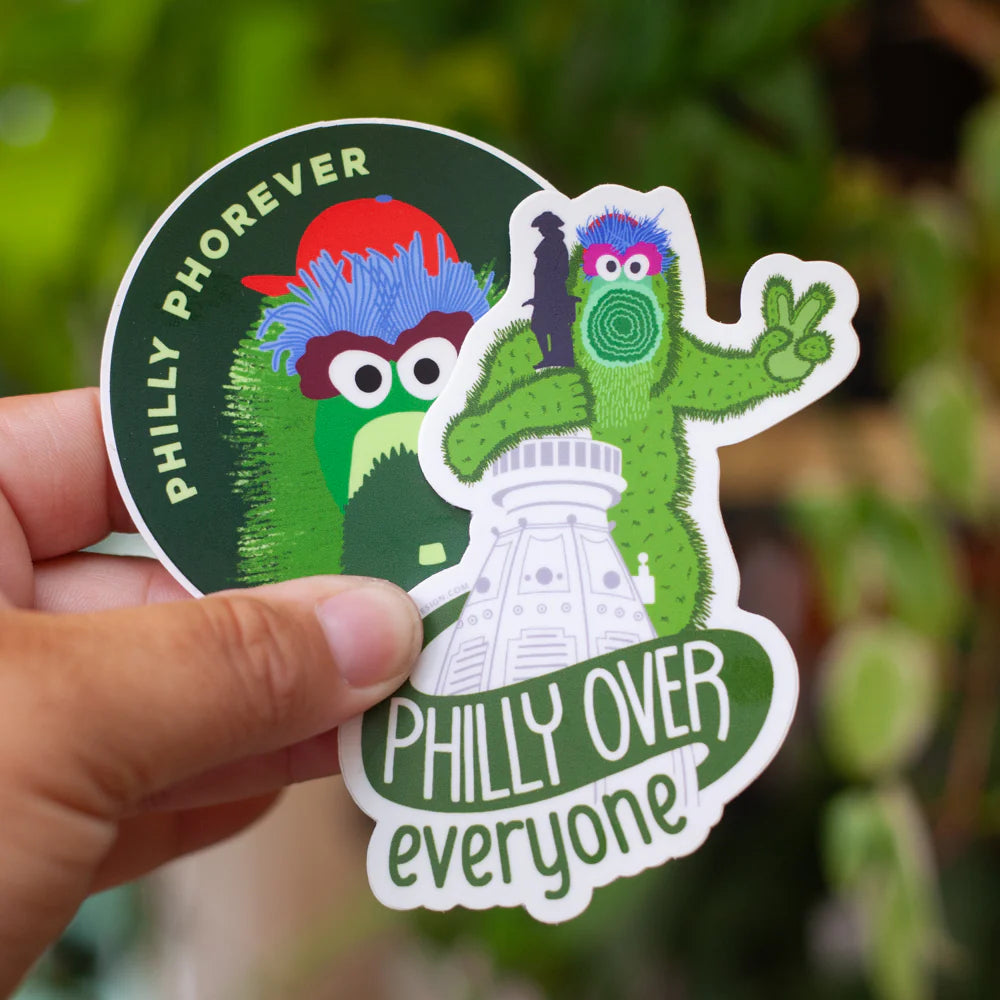 Hand holding a sticker with cartoon characters and text 'PHILLY PHOREVER' and 'PHILLY OVER everyone' against a blurred green background.